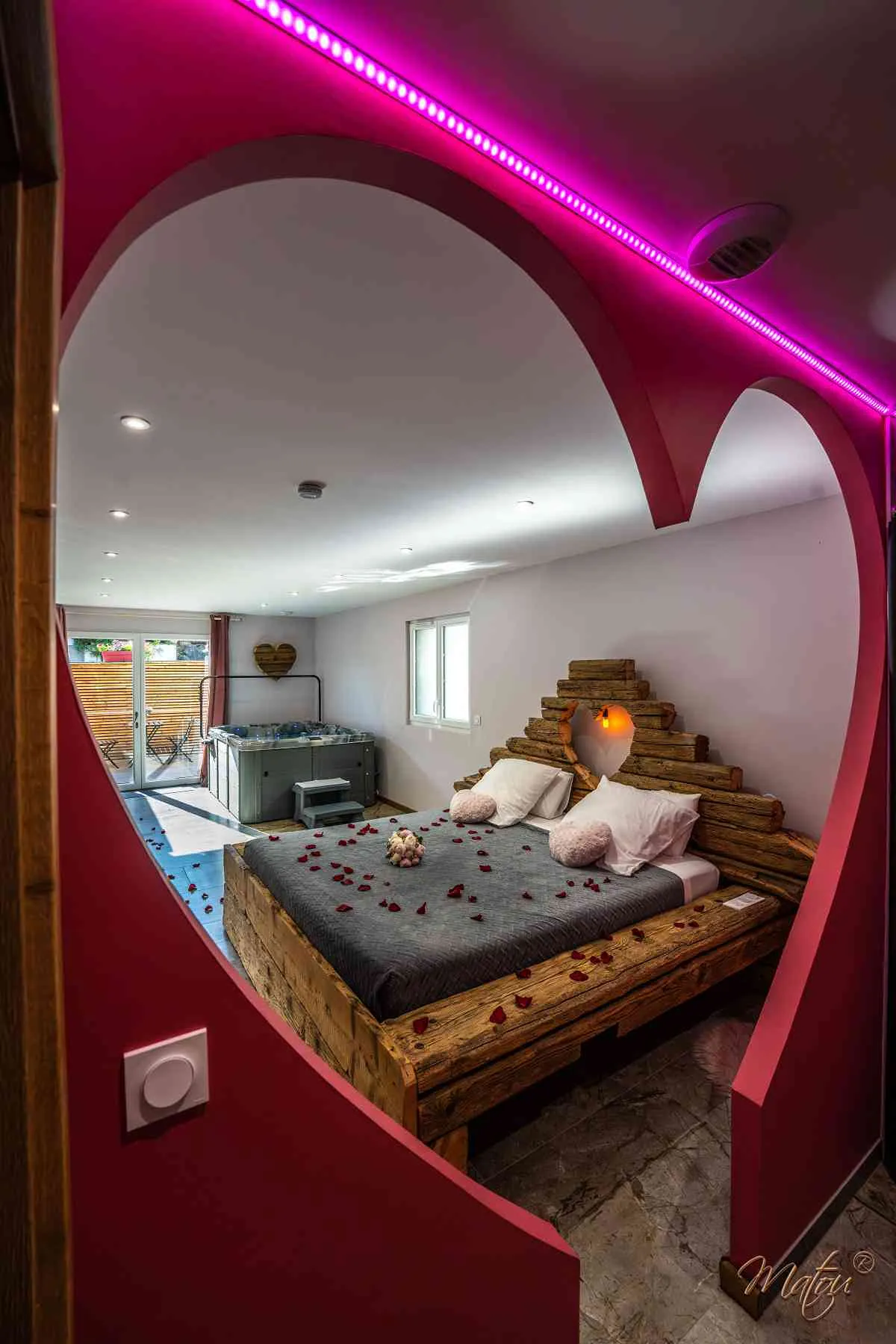 Vue à travers une ouverture en forme de cœur sur le lit et le jacuzzi de la love room Bisou Sucré